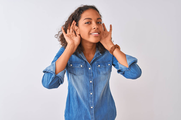 Mujer brasileña joven con camisa de mezclilla de pie sobre fondo blanco aislado tratando de escuchar ambas manos en el gesto de la oreja, curioso por chismes. Problema de audición, sordos
 - Foto, imagen