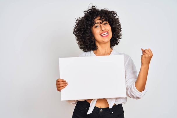 Young arab woman with curly hair holding banner over isolated white background screaming proud and celebrating victory and success very excited, cheering emotion - Foto, Imagen