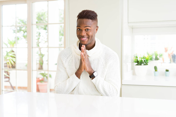 Bello afroamericano sul tavolo bianco che prega con le mani unite chiedendo perdono sorridendo fiducioso
. - Foto, immagini