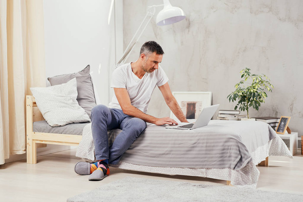 guapo caucásico de mediana edad hombre vestido casual usando el ordenador portátil mientras está sentado en la cama en el dormitorio
. - Foto, Imagen