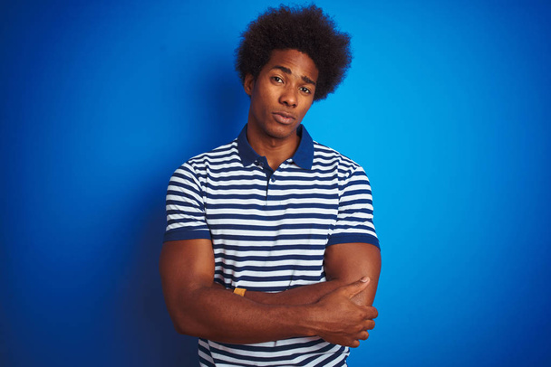 Homme afro-américain aux cheveux afro portant un polo rayé debout sur un fond bleu isolé sceptique et nerveux, désapprouvant l'expression sur le visage avec les bras croisés. Personne négative
. - Photo, image