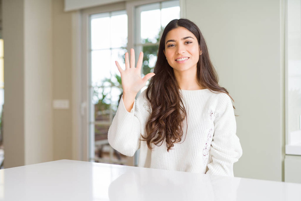 Young beautiful woman at home on white table showing and pointing up with fingers number five while smiling confident and happy. - Foto, afbeelding