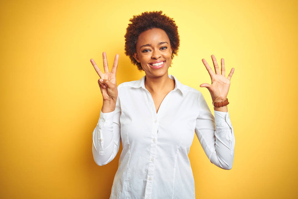 孤立した黄色の背景の上にアフリカ系アメリカ人のビジネスウーマンは、自信と幸せに微笑みながら、指番号8を示し、指を指差す. - 写真・画像