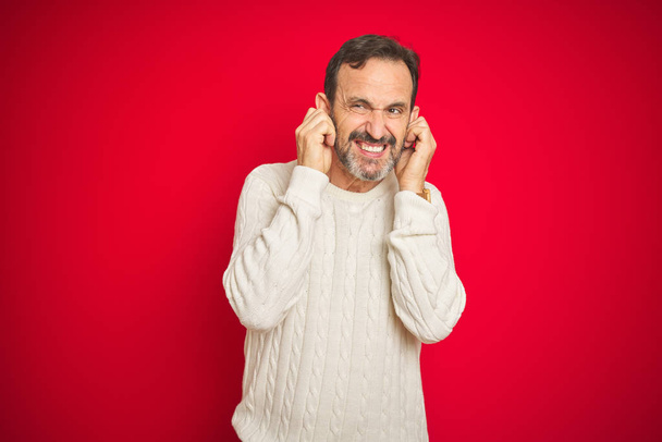 Knappe middelbare leeftijd Senior man met grijze haren over geïsoleerde rode achtergrond bedekken oren met vingers met geïrriteerde uitdrukking voor het lawaai van luide muziek. Doof concept. - Foto, afbeelding