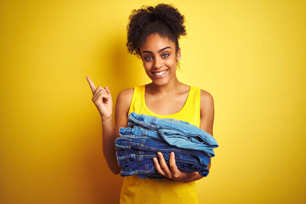Young african american woman holding stack of jeans over isolated yellow background very happy pointing with hand and finger to the side - Foto, imagen