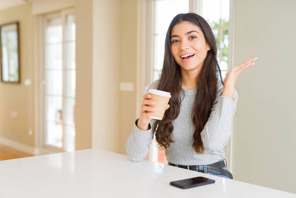 Nuori nainen juo kupin kahvia kotona erittäin onnellinen ja innoissaan, voittaja ilme juhlii voittoa huutaen iso hymy ja nosti kätensä
 - Valokuva, kuva