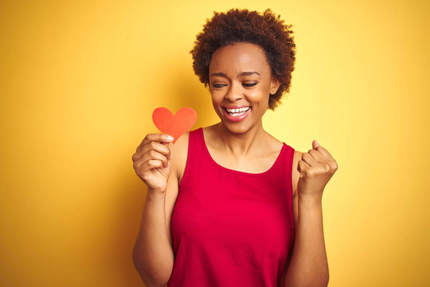 African american woman holding romantic paper hearts over yellow isolated background screaming proud and celebrating victory and success very excited, cheering emotion - Fotoğraf, Görsel