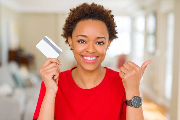 Молодая африканская американка, держащая кредитку, указывая пальцем вверх в сторону, со счастливым лицом, улыбающимся
 - Фото, изображение