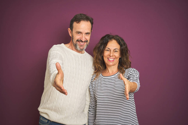 Beautiful middle age couple wearing winter sweater over isolated purple background smiling friendly offering handshake as greeting and welcoming. Successful business. - Foto, Imagem