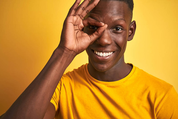 Homme afro-américain portant un t-shirt décontracté debout sur fond jaune isolé avec un visage heureux souriant faisant signe ok avec la main sur les yeux regardant à travers les doigts
 - Photo, image
