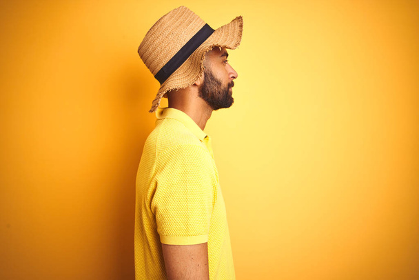 junger indischer Mann im Urlaub mit Sommermütze vor isoliertem gelben Hintergrund zur Seite schauend, entspannte Profilpose mit natürlichem Gesicht mit selbstbewusstem Lächeln. - Foto, Bild