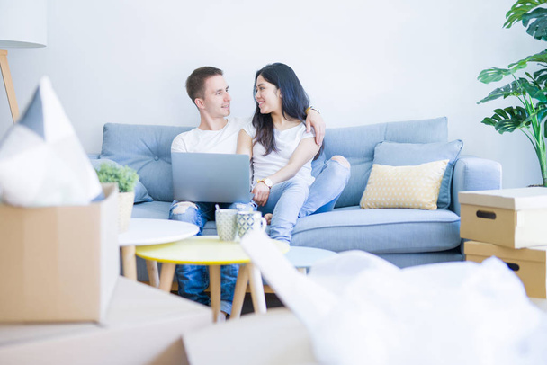 Joven hermosa pareja sentada en el sofá utilizando el ordenador portátil en el nuevo hogar alrededor de cajas de cartón
 - Foto, imagen