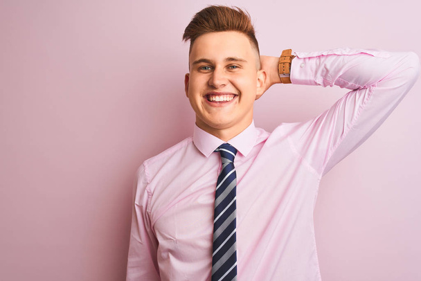 シャツとネクタイを着た若いハンサムなビジネスマンは、手を上げるジェスチャーで自信を持って髪に触れ、魅力的でファッショナブルなポーズをとる孤立したピンクの背景の上に立って - 写真・画像
