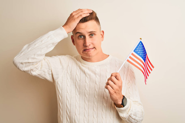 Joven hombre guapo sosteniendo la bandera de EE.UU. sobre fondo blanco aislado estresado con la mano en la cabeza, conmocionado con vergüenza y cara sorpresa, enojado y frustrado. Miedo y malestar por error
. - Foto, Imagen