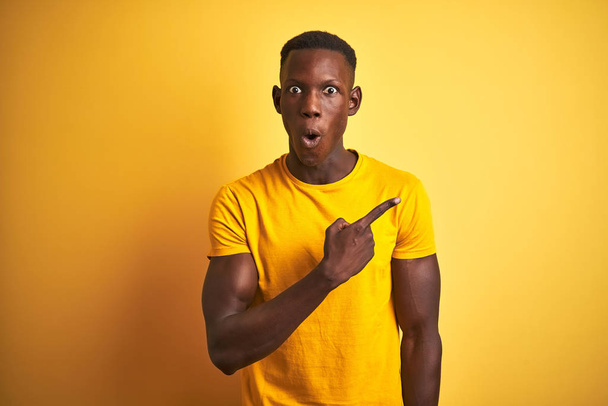 Fiatal afro-amerikai férfi visel alkalmi póló állt át elszigetelt sárga háttér meglepett mutatva ujját az oldalra, nyitott száj csodálkozva kifejezés. - Fotó, kép