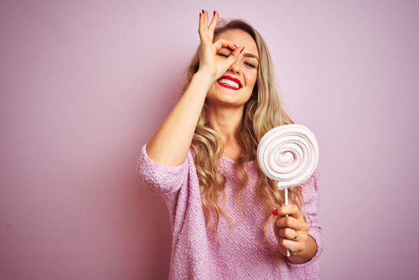 Jovem mulher bonita comendo doces doces sobre fundo isolado rosa com o rosto feliz sorrindo fazendo sinal ok com a mão no olho olhando através dos dedos
 - Foto, Imagem