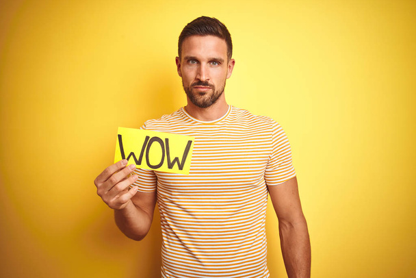Young man holding wow text banner over yellow isolated background with a confident expression on smart face thinking serious - Φωτογραφία, εικόνα