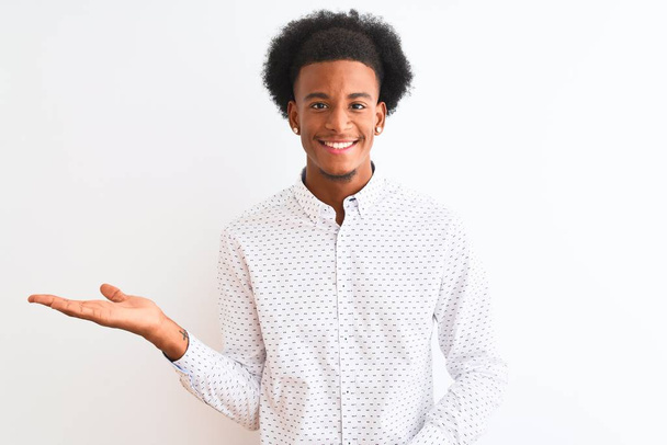 孤立した白い背景の上に立つエレガントなシャツを着た若いアフリカ系アメリカ人男性は、カメラを見て手のひらを指差して陽気な提示を笑顔にする. - 写真・画像