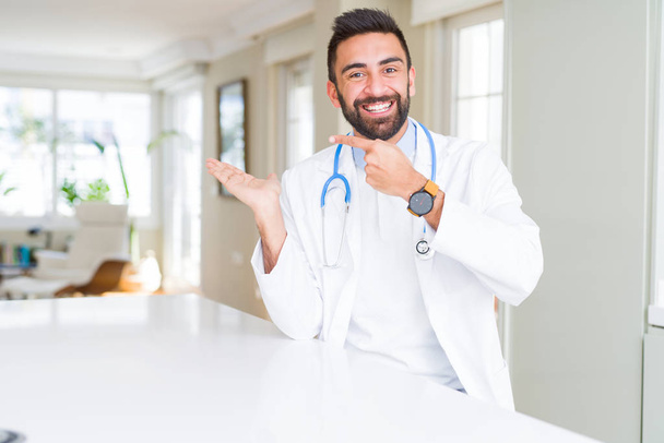 Beau médecin hispanique homme portant stéthoscope à la clinique étonné et souriant à la caméra tout en présentant avec la main et pointant du doigt
. - Photo, image