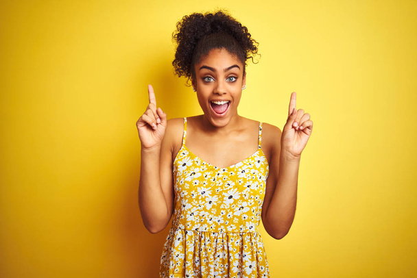 African American kobieta sobie casual kwiatowy strój stojący na białym tle żółte uśmiechnięty zdziwiony i zaskoczony i wskazując na palce i podniesione ramiona. - Zdjęcie, obraz