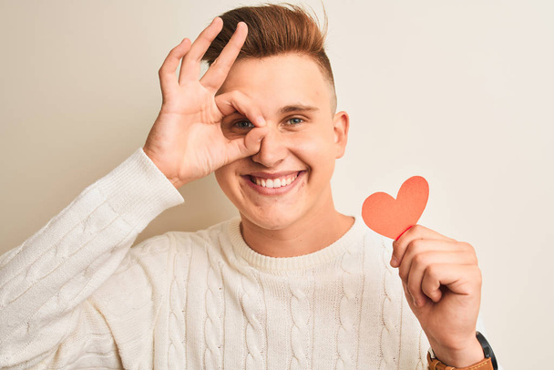Young handsome romantic man holding paper hearts standing over isolated white background with happy face smiling doing ok sign with hand on eye looking through fingers - Φωτογραφία, εικόνα