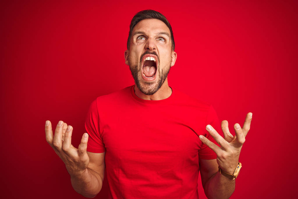 Jonge knappe man dragen casual t-shirt over rode geïsoleerde achtergrond gek en boos schreeuwen en schreeuwen met agressieve expressie en armen verhoogd. Frustratie concept. - Foto, afbeelding