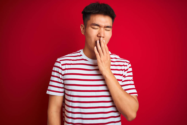 Joven asiático chino hombre usando rayas camiseta de pie sobre aislado rojo fondo aburrido bostezo cansado cubriendo la boca con la mano. Inquietud y somnolencia
. - Foto, imagen