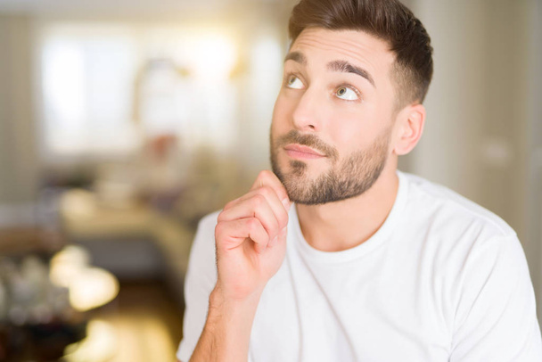 Mladý pohledný muž nosí ležérní bílé tričko doma s rukou na bradě přemýšlet o otázce, zamyšlený výraz. Usmívající se zamyšlenou tváři. Pochybnost koncept. - Fotografie, Obrázek