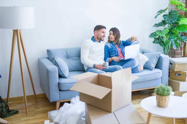 Young beautiful couple sitting on the sofa drinking cup of coffee using laptop at new home around cardboard boxes - Photo, Image