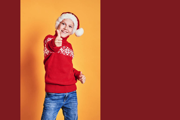 Boy in Santa hat showing thumbs up on yellow - Foto, immagini