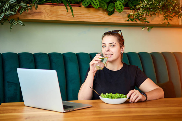 Jovem mulher feliz bebe água no belo interior com flores verdes no fundo e ingredientes frescos na mesa. Conceito de alimentação saudável
 - Foto, Imagem
