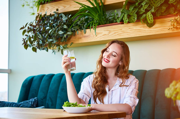 Young happy woman drinks water in the beautiful interior with green flowers on the background and fresh ingredients on the table. Healthy food concept - Photo, Image