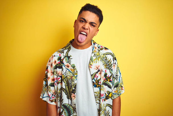 Young brazilian man on vacation wearing summer floral shirt over isolated yellow background sticking tongue out happy with funny expression. Emotion concept. - Fotoğraf, Görsel