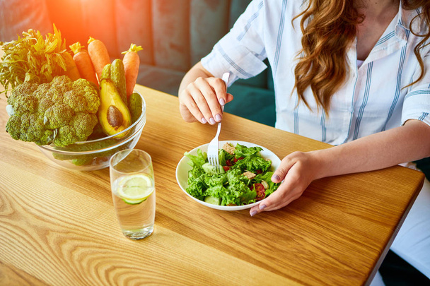 背景に緑の花とテーブルの上に新鮮な食材と美しいインテリアでサラダを食べる若い幸せな女性。健康食品のコンセプト - 写真・画像