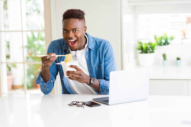 Hermoso joven africano hombre de negocios comiendo comida asiática a domicilio y trabajando con computadora, disfrutando fideos sonriendo
 - Foto, imagen