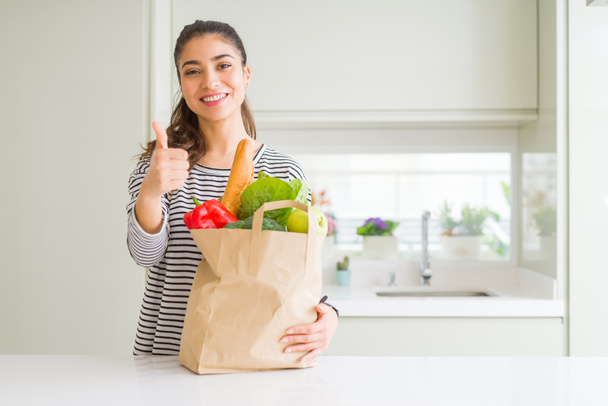 Young woman holding paper bag full of groceries happy with big smile doing ok sign, thumb up with fingers, excellent sign - Foto, afbeelding