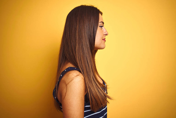 Młoda piękna kobieta w paski t-shirt stoi na odizolowanym żółtym tle patrząc na bok, relaks profil pozować z naturalną twarz z pewnym uśmiechem. - Zdjęcie, obraz
