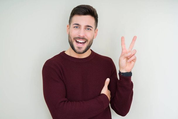 Young handsome man wearing a sweater over isolated background smiling with happy face winking at the camera doing victory sign. Number two. - Fotó, kép