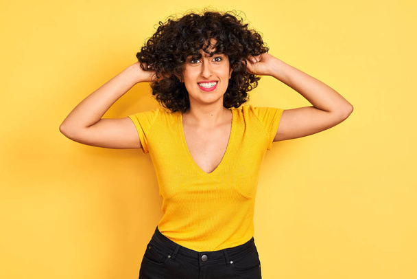 Jeune femme arabe aux cheveux bouclés portant un t-shirt debout sur fond jaune isolé Souriant tirant les oreilles avec les doigts, geste drôle. Problème d'audition
 - Photo, image