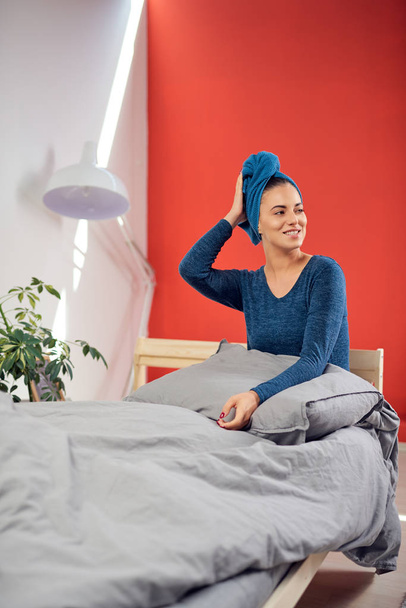 Beautiful smiling Caucasian woman with blue towel on head sitting on the bed in bedroomm in the morning. - Photo, Image