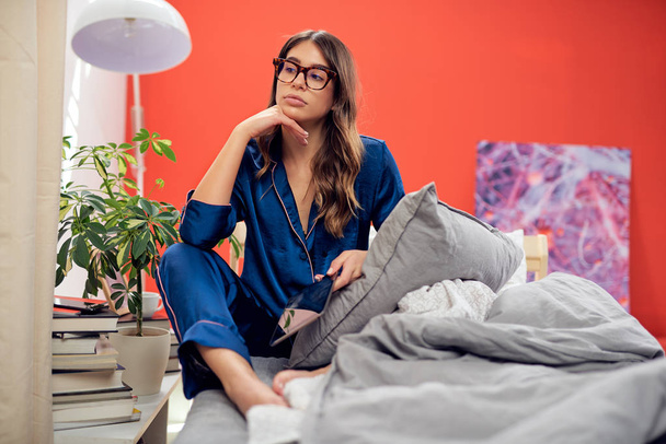 Πανέμορφο Καυκάσιος μελαχρινή με γυαλιά και ντυμένος με μπλε πιτζάμες κάθεται στο κρεβάτι στο υπνοδωμάτιο, το πρωί και χρησιμοποιώντας tablet. - Φωτογραφία, εικόνα