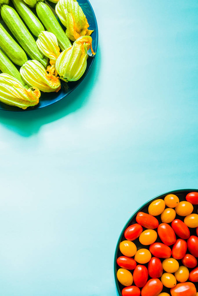 frische rohe Zucchini (Zucchini) mit Blüten und Kürbisblüten und kirschfarbenen Tomaten auf blauem Teller vor blauem Hintergrund, Draufsicht, natürliches Licht. - Foto, Bild