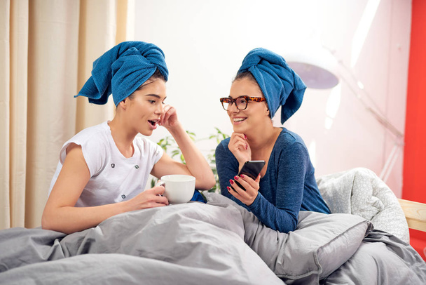 Twee aantrekkelijke vriendinnen zittend op het bed in de ochtend met handdoeken op de kop, kijkend naar slimme telefoon en koffie drinken. - Foto, afbeelding