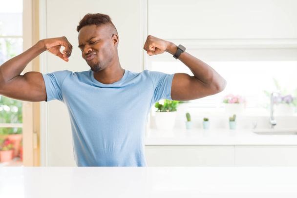 Симпатичный африканский американец в обычной футболке дома показывает мышцы рук, улыбающиеся с гордостью. Фитнес-концепция
. - Фото, изображение