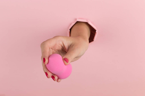 Girl hand holds a beauty blender for makeup from a hole in a pink background, copy space. - Foto, Imagem