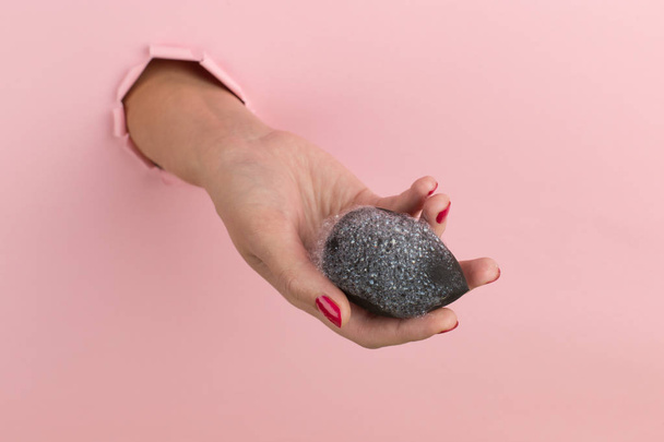 Girl hand holds a beauty blender for makeup from a hole in a pink background, copy space. Sponge in soapy foam, how to clean. - Foto, Bild