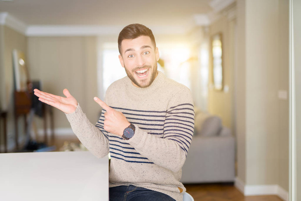 Młody przystojny mężczyzna ubrany w domu sweter zdumiony i uśmiechnięty do aparatu podczas prezentacji z ręki i wskazując palcem. - Zdjęcie, obraz