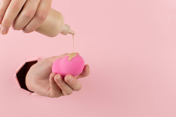 Girl hand holds a beauty blender in foundation, concealer from a hole in a pink background. Makeup artist concept, copy space. - Φωτογραφία, εικόνα