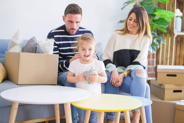 Beautiful family, parents sitting on the sofa drinking coffee looking his kid playing at new home around cardboard boxes - Фото, изображение