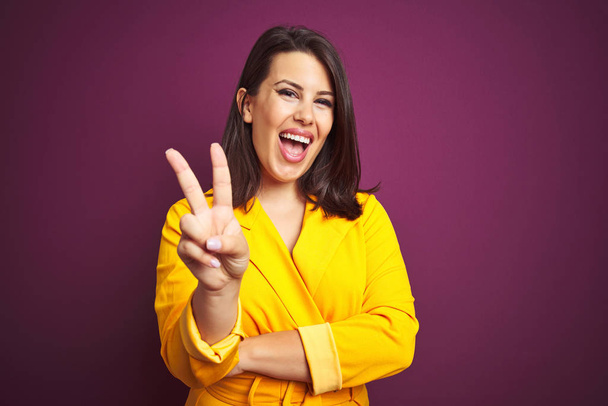 Young beautiful brunette woman wearing elegant yellow jacket over purple isolated background smiling with happy face winking at the camera doing victory sign. Number two. - Fotografie, Obrázek